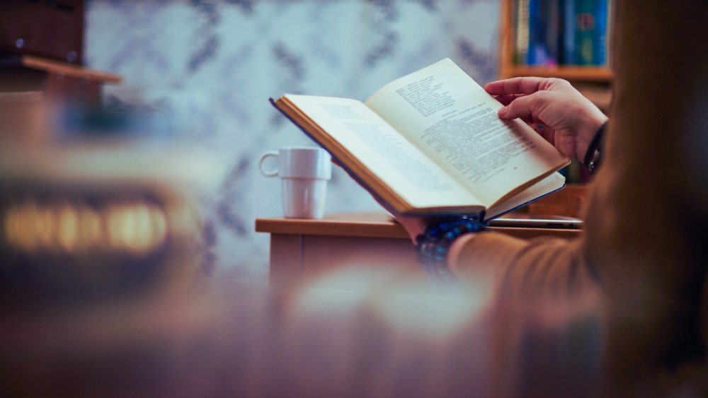 Une femme assise dans une bibliothèque tient un livre ouvert devant elle. Une tasse de café se trouve sur la table à laquelle la lectrice est accoudée.