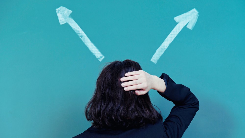 Femme de dos en proie à l'incertitude. Elle pose une main sur sa tête et regarde deux flèches blanches tracées sur le mur bleu en face d'elle. Elle incarne l'indécision et le non-choix.